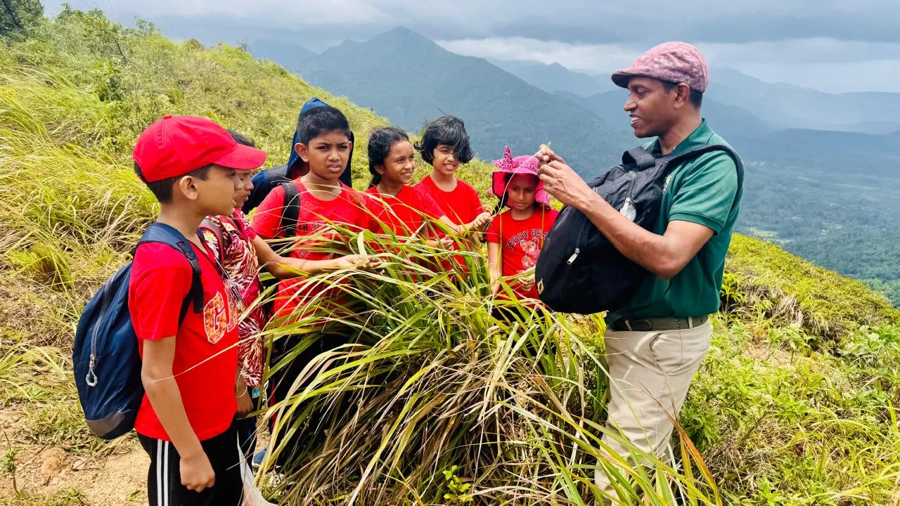 Nayana Pradeep showing a plant to students on a hillside, with mountains in the background.