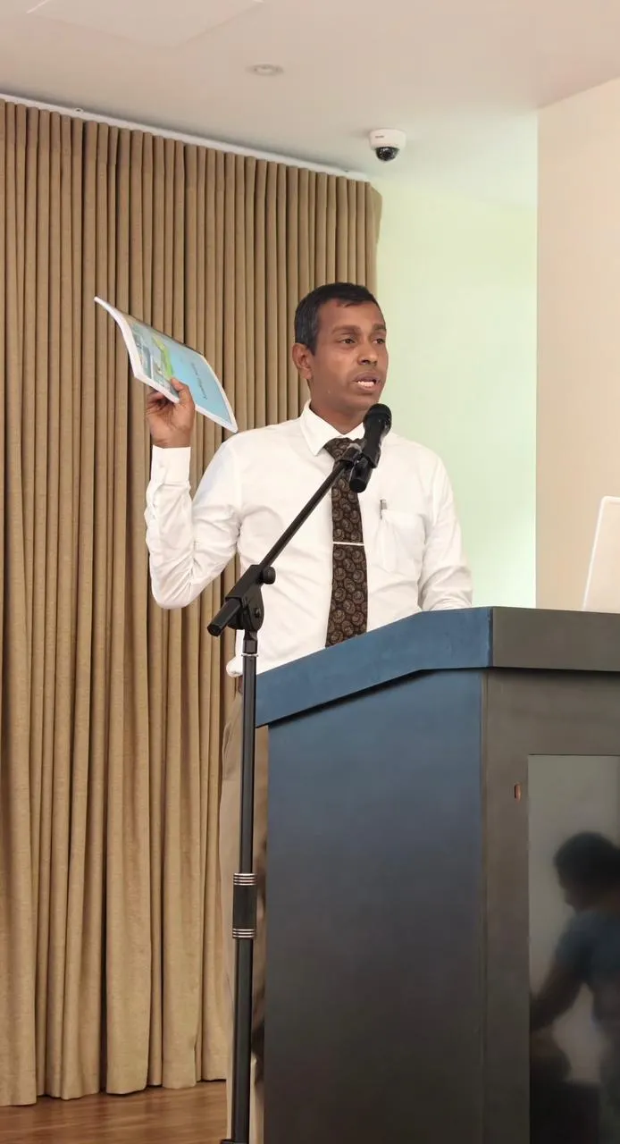 Nayana Pradeep addressing school principals from the podium, holding up one of his books.
