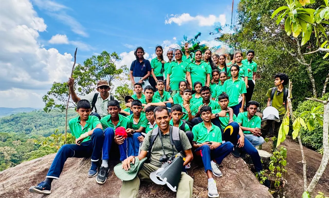 Group photograph of the Vidulipura Maha Vidyalaya Exploration & Research Society on a rock outcrop overlooking the forest, with Nayana Pradeep seated at the front.