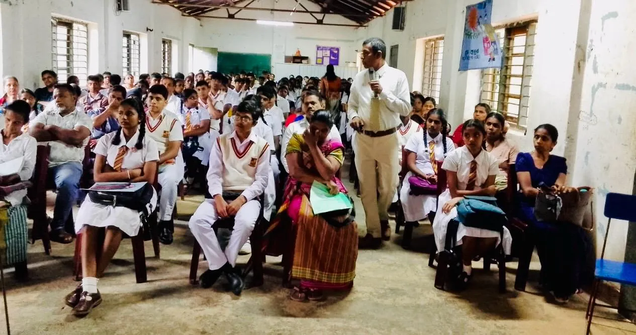 Nayana Pradeep speaking with a microphone while standing among Advanced Level students and parents in the Ambagamuwa Central College hall.