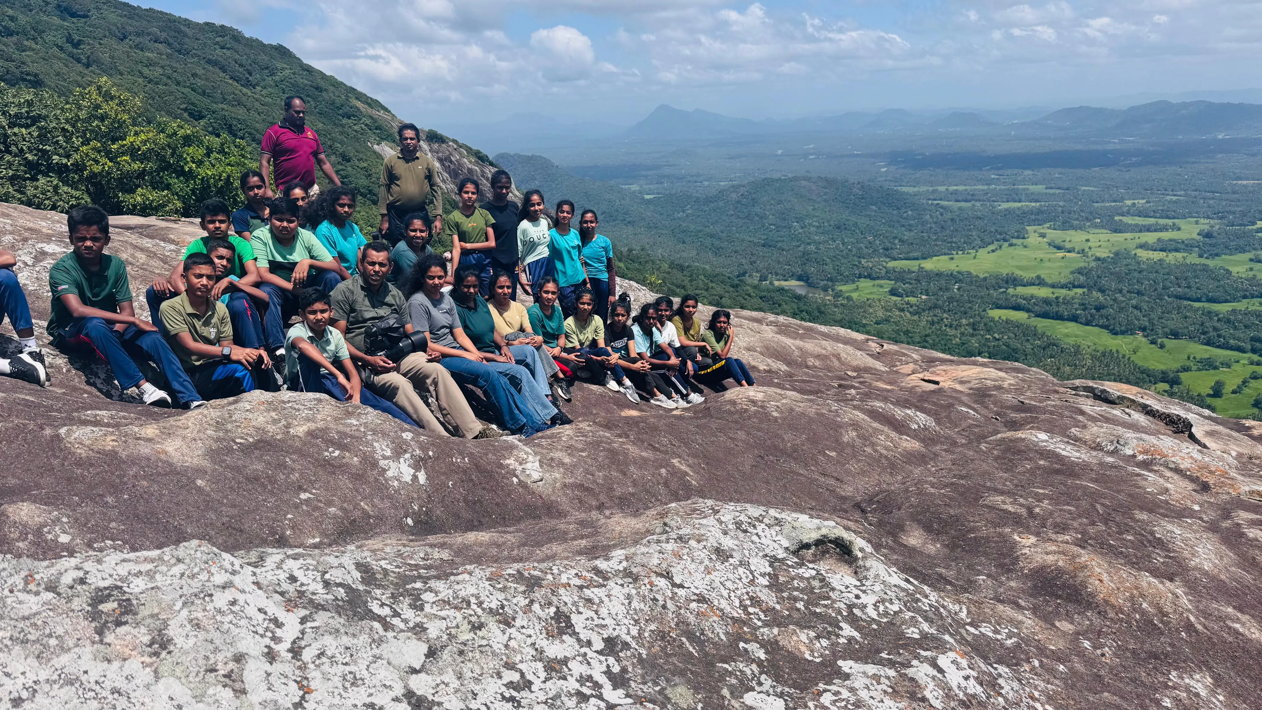 Students of Ambagamuwa Maha Vidyalaya with Nayana Pradeep on the summit rock of Dolukanda, overlooking a sweeping valley.