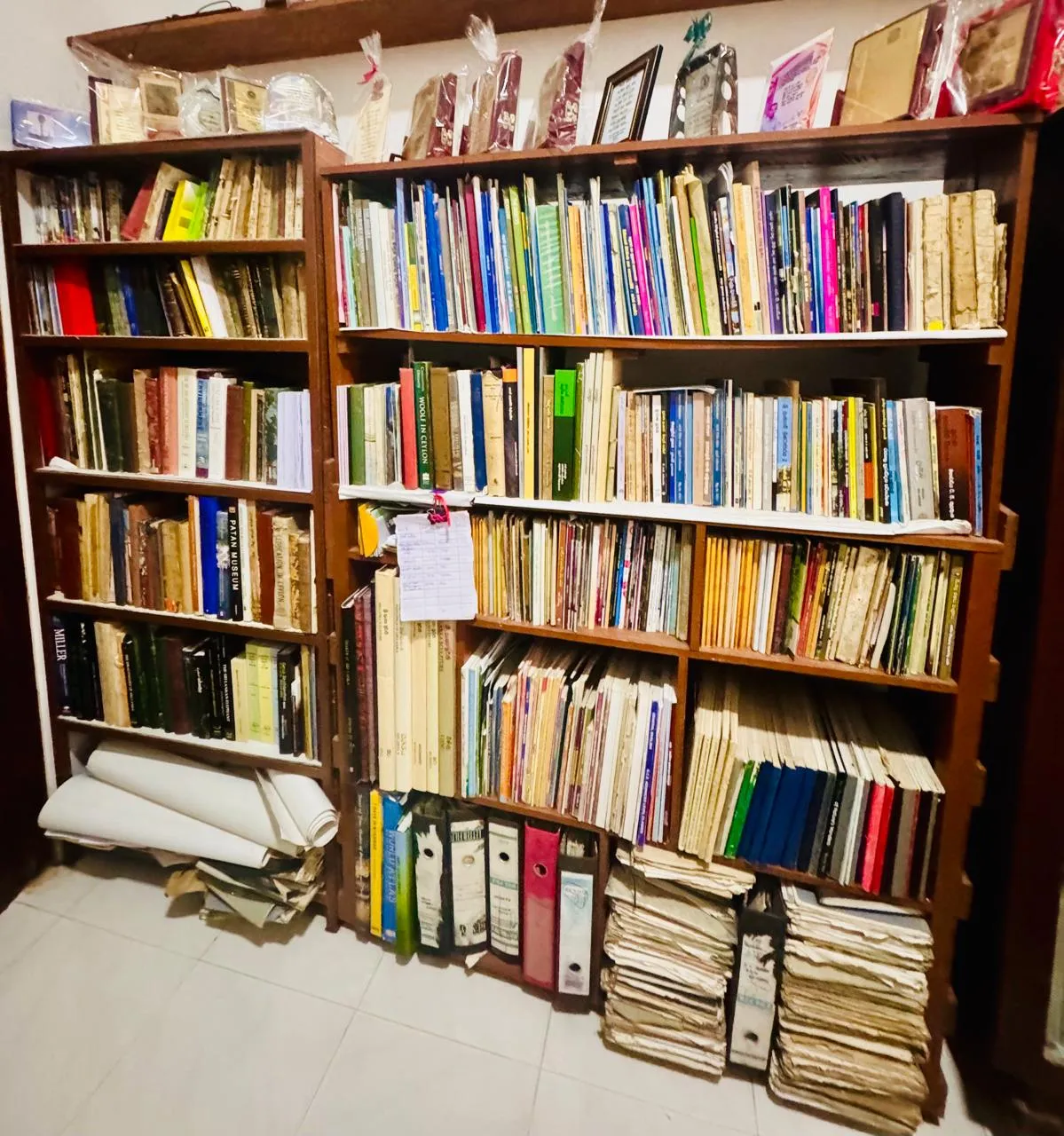 A wall of colourful book spines above stacked bound journals and paper archives.