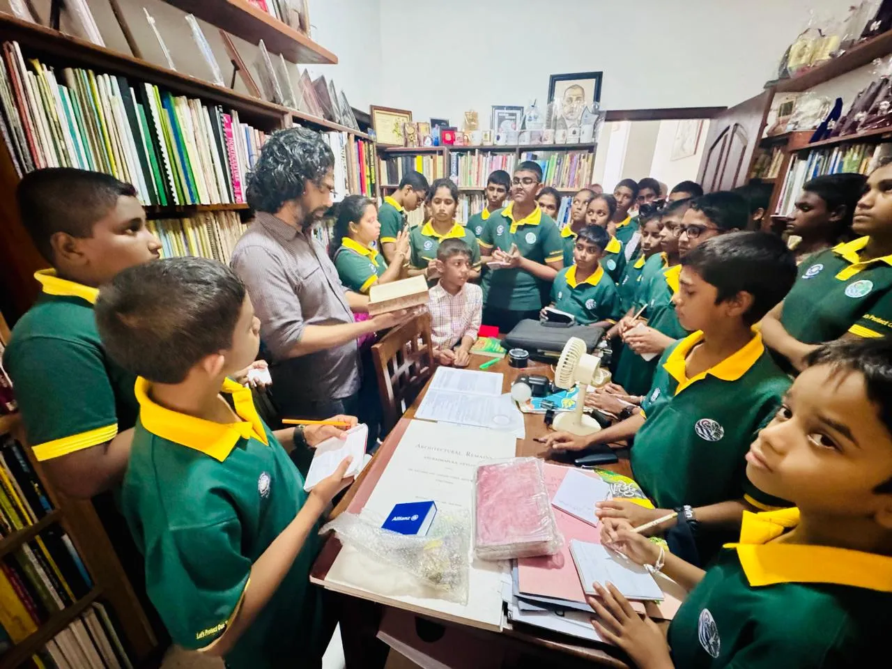 A group of schoolchildren in green and yellow uniforms crowded around a desk in the library, taking notes while Nayana shows them an old bound volume.